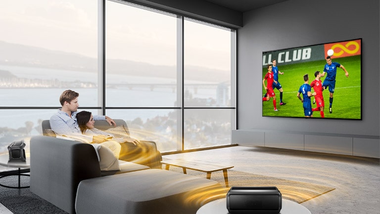 رجل وامرأة يشاهدان الألعاب الرياضية على شاشة التلفزيون في غرفة المعيشة مع مكبرات صوت خلفية تعمل بتقنية البلوتوث