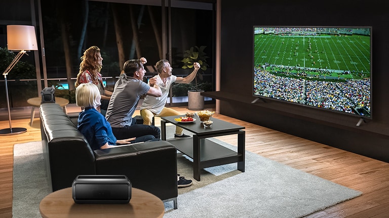 أناس يشاهدون الألعاب الرياضية على شاشة التلفزيون في غرفة المعيشة مع مكبرات صوت خلفية تعمل بتقنية البلوتوث.