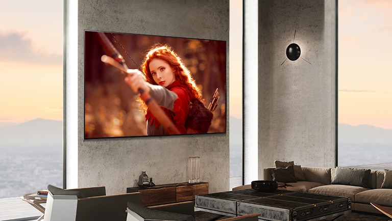 تلفاز كبير مُثبَّت على جدار حجري رمادي بجوار نوافذ ممتدة من الأرض حتى السقف في غرفة حديثة.  تظهر على الشاشة امرأة ذات شعر أحمر تحمل قوسًا موجهًا نحو الكاميرا. 