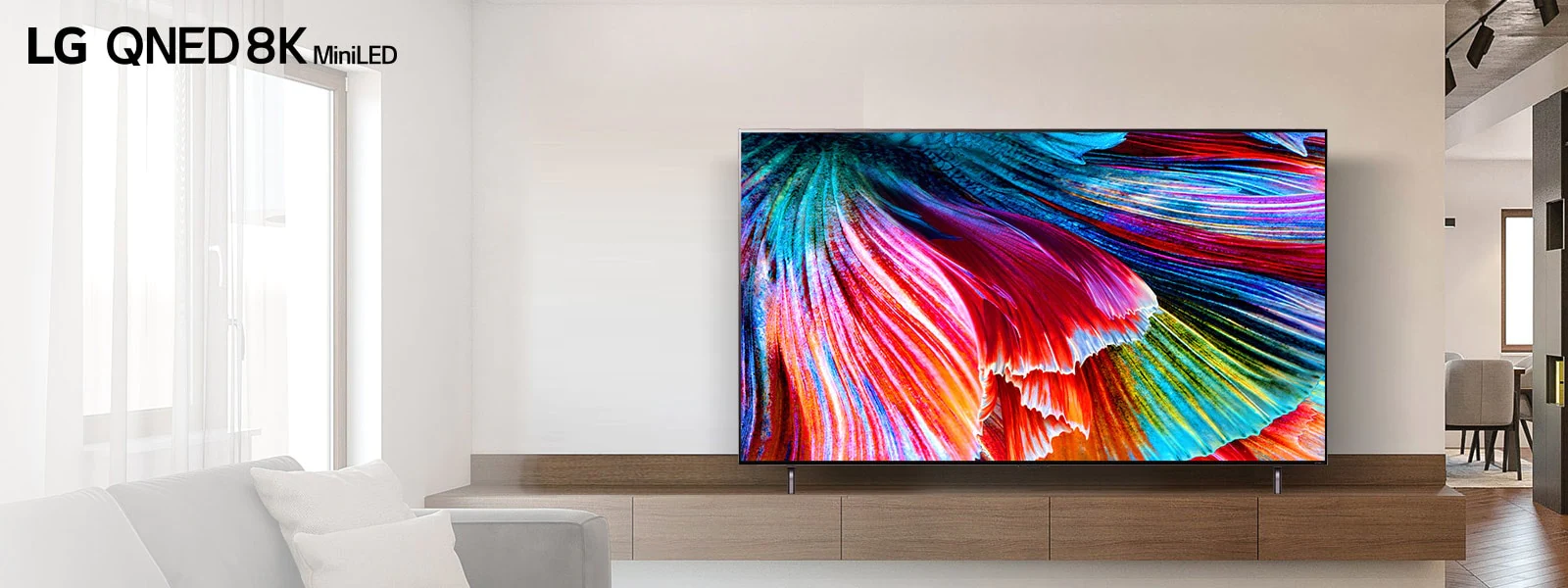 يقف تلفزيون QNED MiniLED على خزانة خشبية مقابل الجدار الخفيف في تصميم داخلي حديث فاتح. تعرض الشاشة صورة مقربة لبتلات متعددة الألوان تتصاعد.