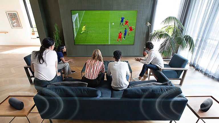 مشهد يوضح تجمع مكون من 5 أشخاص أمام شاشة تلفزيون مسطحة مثبتة على الجدار لمشاهدة إحدى مباريات كرة القدم.