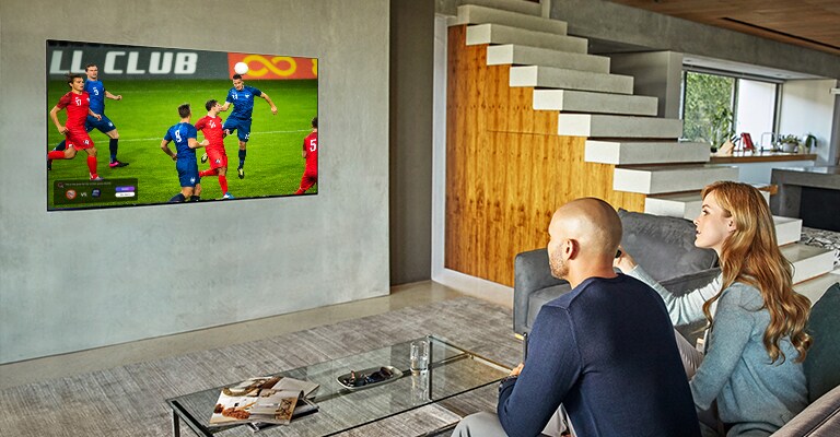صورة يظهر بها رجل وامرأة يجلسان خلف طاولة قهوة أمام تلفزيون مثبت على الجدار يشاهدان إحدى مباريات كرة القدم.