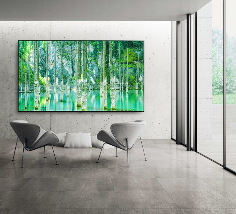 صورة توضح شاشة تلفزيون QNED MiniLED كبيرة الحجم من إل جي مثبتة على جدار رمادي من الخرسانة المجردة. شاشة تعرض مشهدًا طبيعيًا أخضر اللون مع وجود كرسيين أمام التلفزيون.