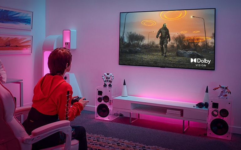 صورة توضح فتاة تلعب لعبة تقمص الأدوار بجهاز تحكم وشاشة تلفزيون كبيرة معلقة على الجدار في غرفتها الوردية.