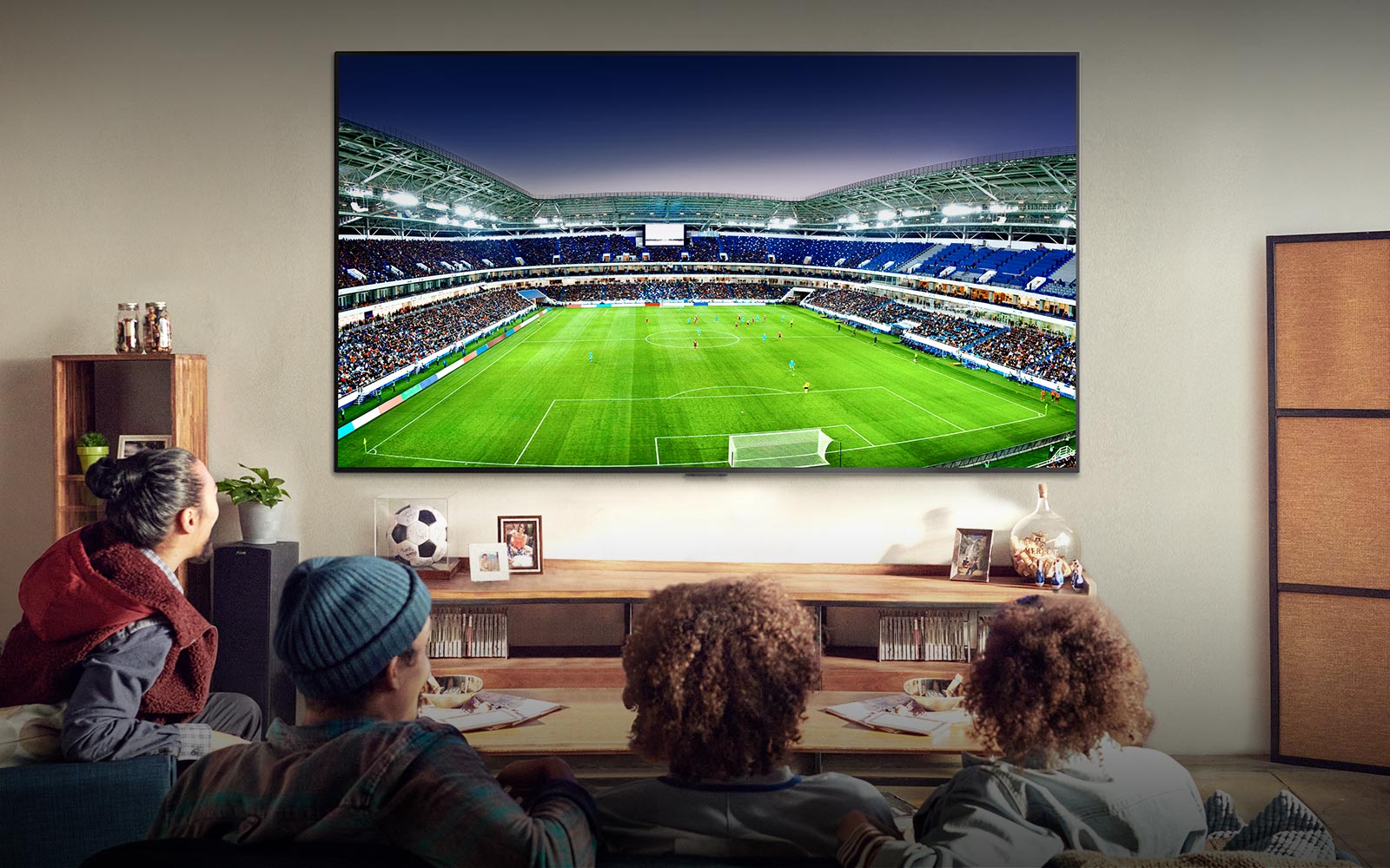 Four people sitting on a couch watching a large screen TV displaying a soccer stadium filled with spectators.