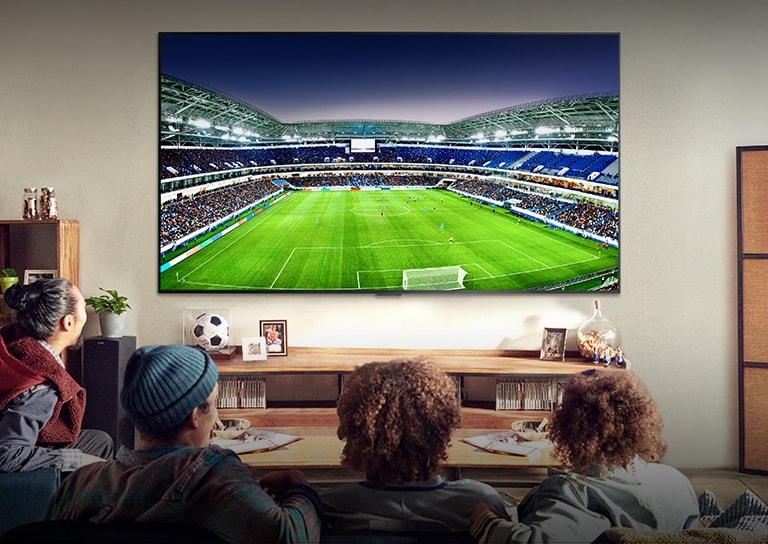 Four people sitting on a couch watching a large screen TV displaying a soccer stadium filled with spectators.