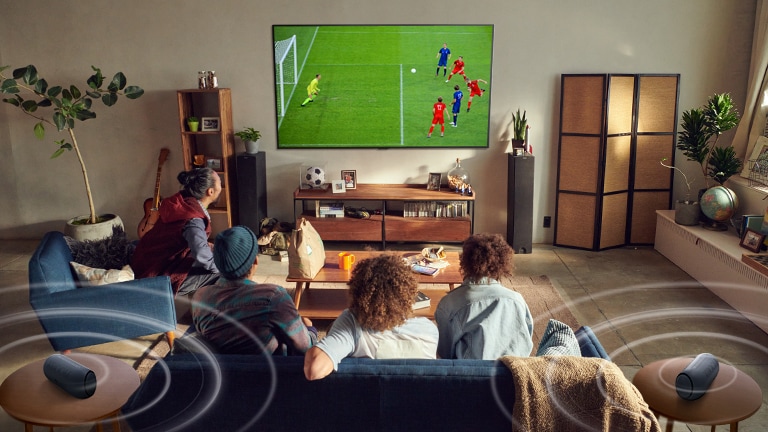 خمسة أشخاص جالسين على الأريكة يشاهدون شاشة تلفاز كبيرة تعرض مباراة كرة قدم مع مكبرات صوت تعمل بتقنية البلوتوث تصدر أصواتًا محيطة. 