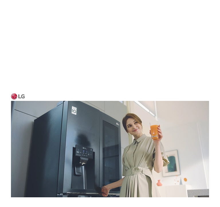 A woman stands in front of the refrigerator holding a glass of juice and smiling as she closes the door.