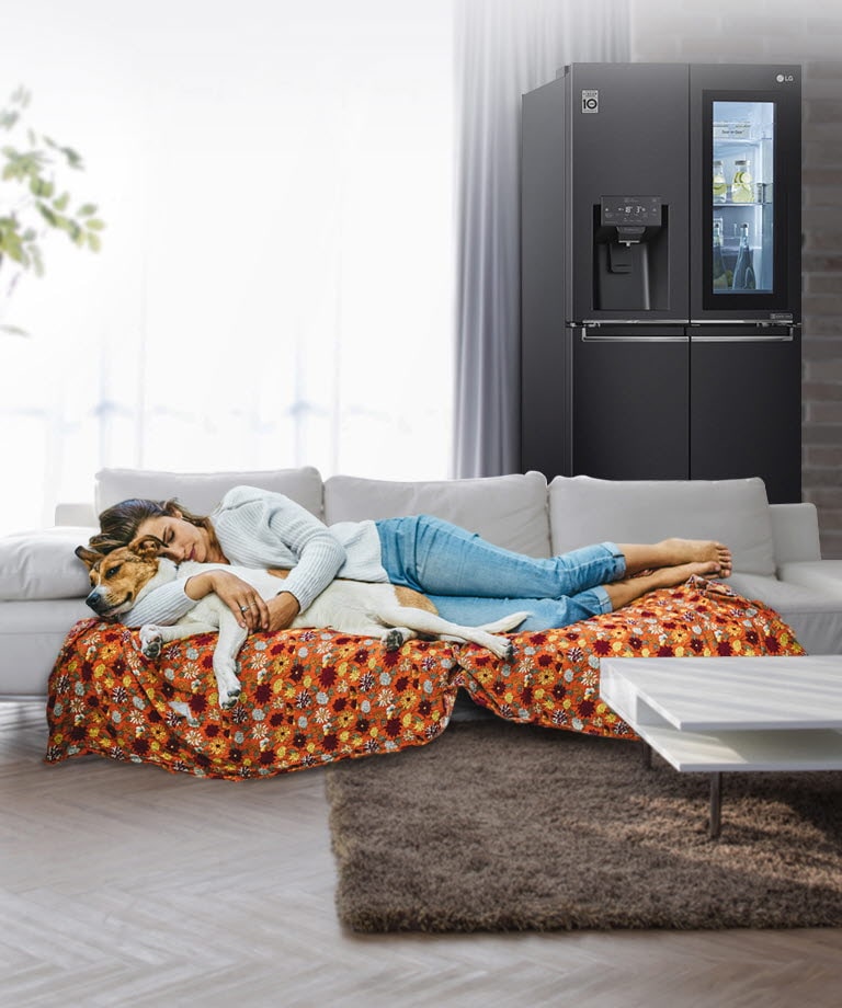 A refrigerator sits against a brick wall with a sofa to the side and a woman and dog sleeping peacefully on the sofa.