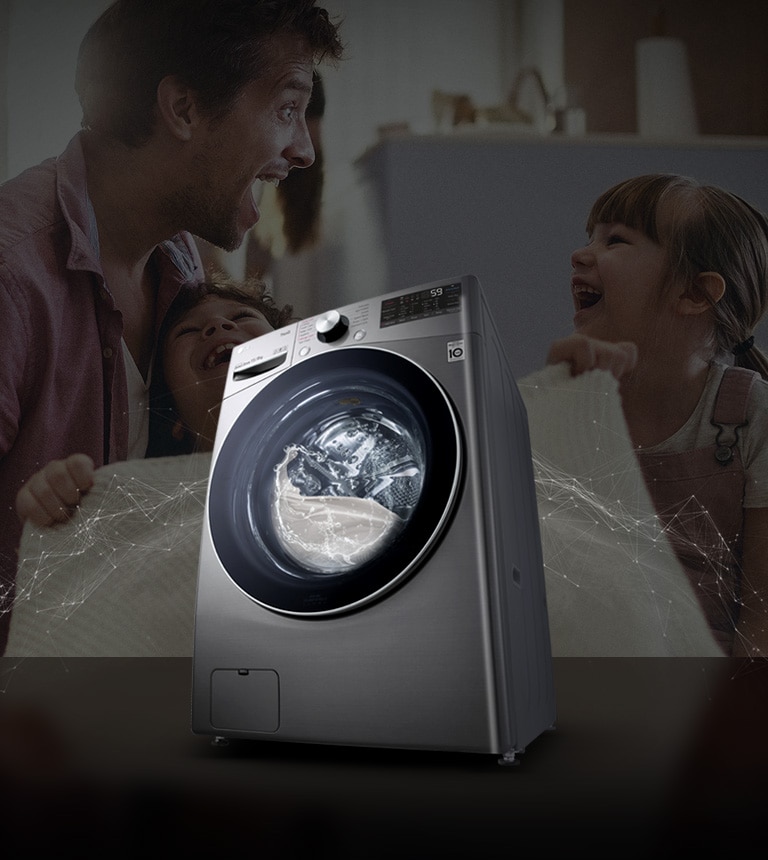 Father and daughters laugh in the background as they hold a clean blanket. A White washing machine front load washer in the foreground.