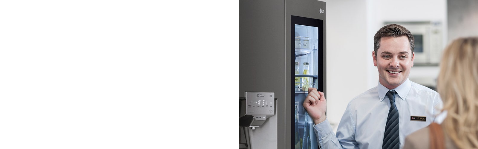 A man in a shirt and tie smiling while indicating a refrigerator behind him as he speaks to a woman with her back to the camera.