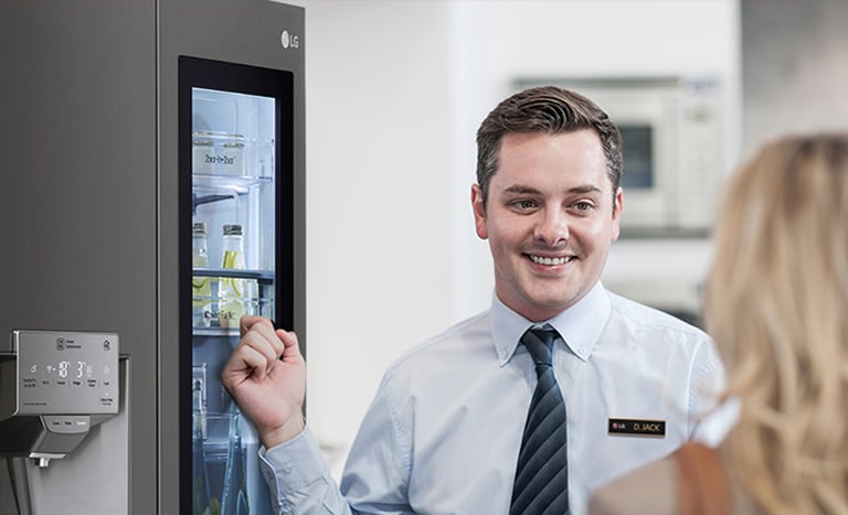 A man in a shirt and tie smiling while indicating a refrigerator behind him as he speaks to a woman with her back to the camera.