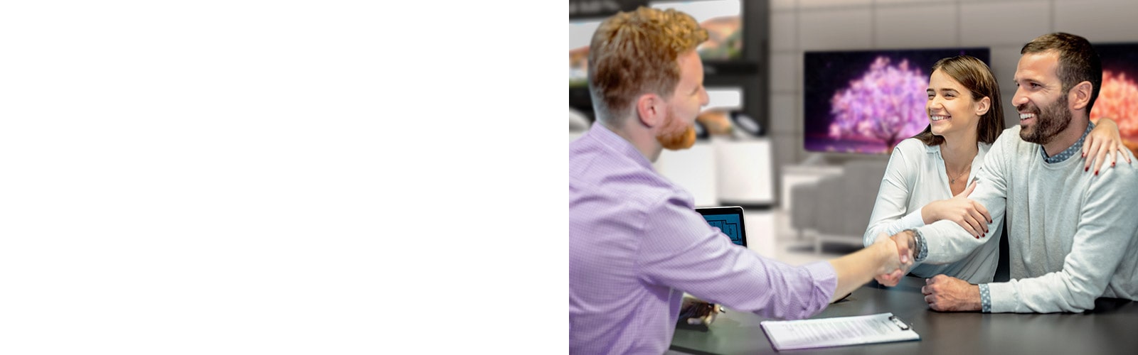 A man in a purple shirt shakes hands with a couple while sat at a table in front of several display unit TVs.