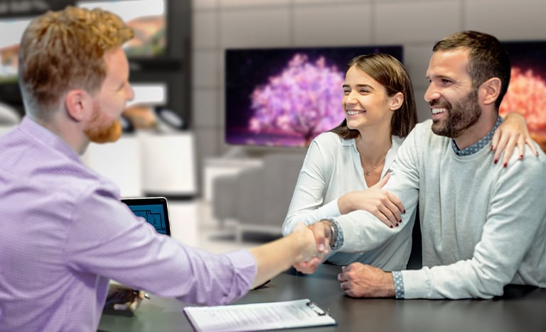 A man in a purple shirt shakes hands with a couple while sat at a table in front of several display unit TVs.