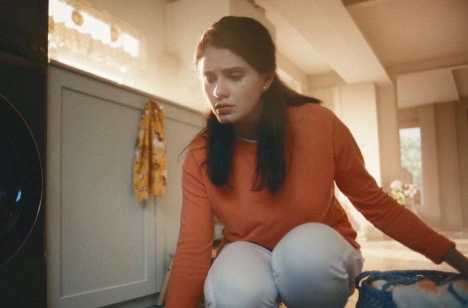 Image shows a woman holding a laundry basket and squatting to touch her kitchen floor.