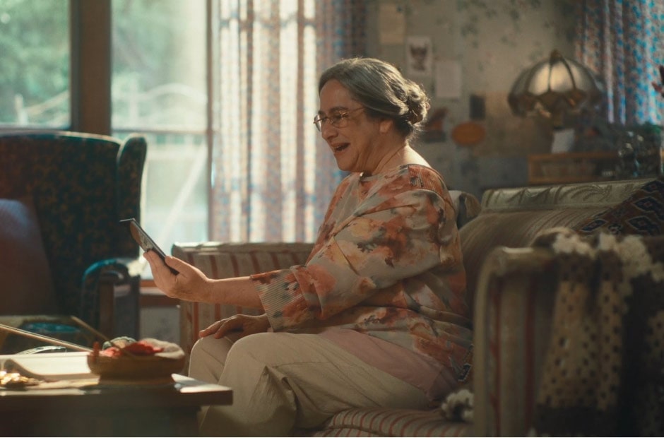 Image shows an older woman sitting in her living room and smiling at a smartphone that’s in her hand.